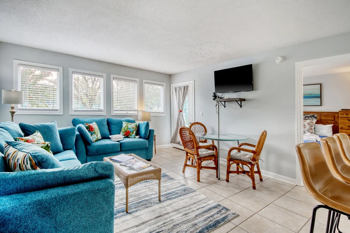 The image shows a bright, cheerful living and dining area featuring plush teal sofas, colorful accent cushions, a sleek glass dining table, and a wall-mounted TV—creating a welcoming space for relaxation and socializing after a day at the beach.