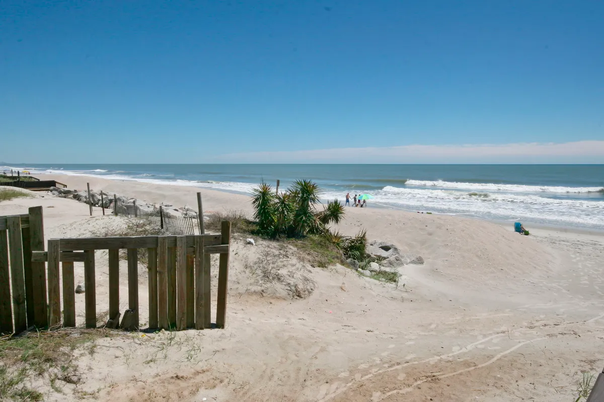A tranquil view of the beach with soft white sand, gentle ocean waves, grassy dunes, and people strolling or playing in the distance. A wooden gate frames the entry onto the sand, inviting guests to step directly onto the beachfront.