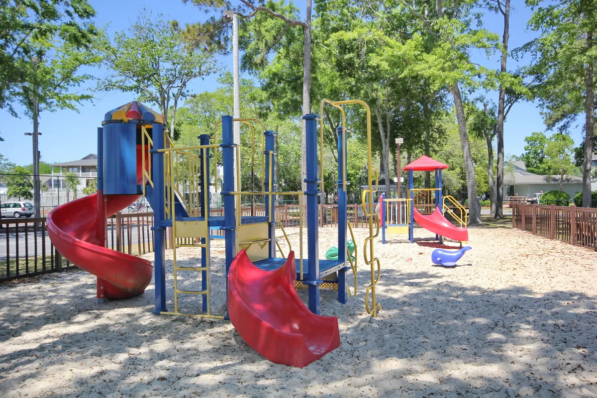 A colorful playground area with sand underfoot and vibrant play structures is framed by surrounding trees, creating an inviting space for families. Children are seen enjoying the equipment, while adults can easily gather nearby for picnics and supervision.
