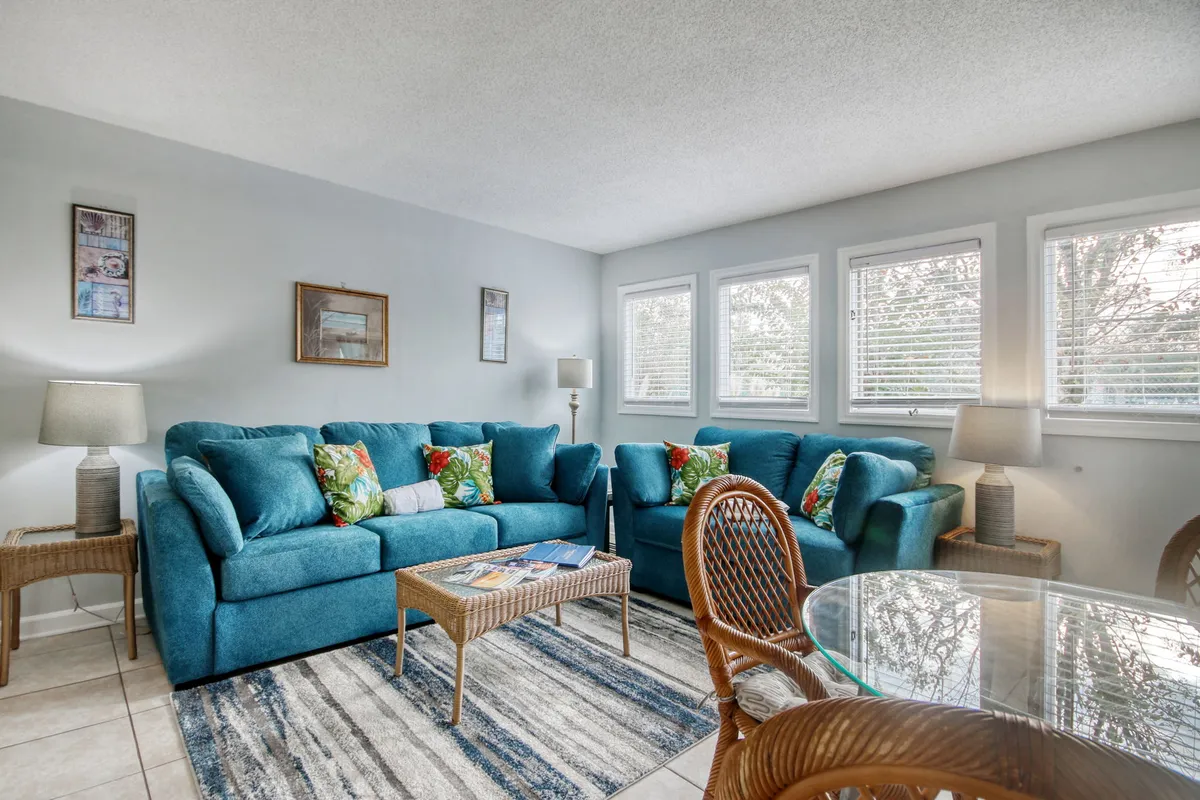 A spacious living room featuring turquoise couches with colorful throw pillows, a glass dining table, and ample natural light. The setting is both stylish and welcoming, ideal for relaxing, socializing, or enjoying a cozy meal at home.
