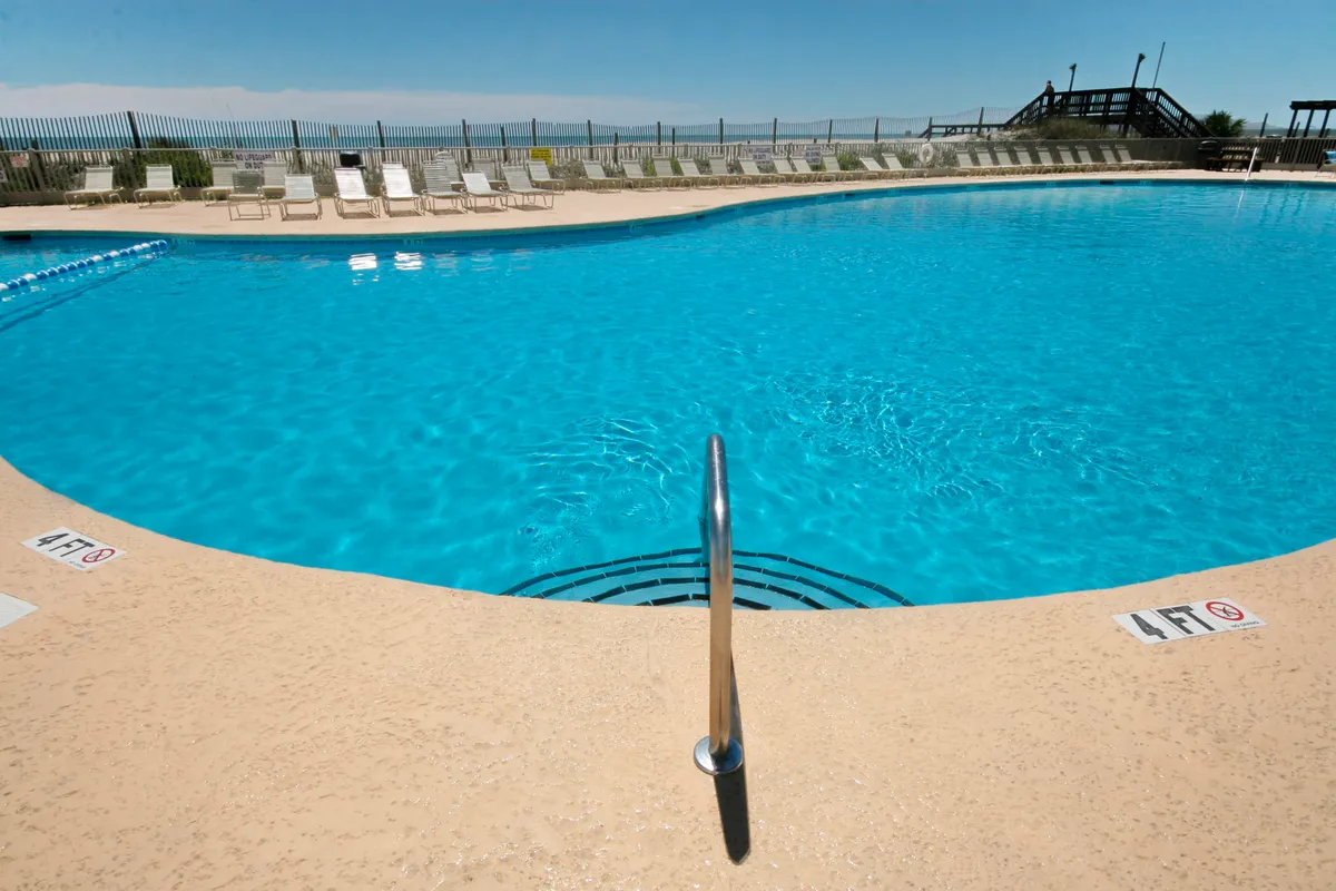 This image showcases a sparkling swimming pool surrounded by poolside loungers and a calm water surface. The pool area is open and sun-kissed, with plenty of space for groups to gather and relax together.