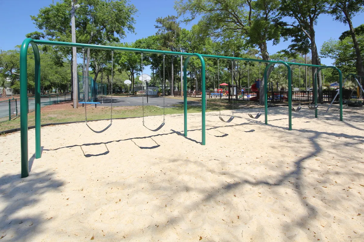 A colorful, well-equipped playground with swings and a modern play structure sits adjacent to a basketball court, surrounded by green grass and trees. Children play and families relax nearby, creating an active and joyful outdoor environment.