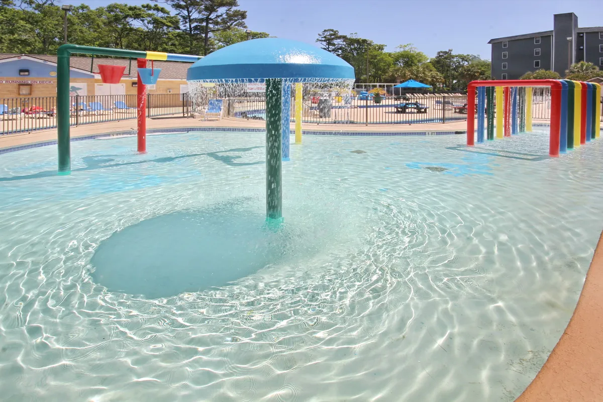 An energetic water play structure rises from the sparkling pool, with children darting through sprays of water and families lounging underneath umbrellas nearby. The scene is colorful, animated, and perfectly captures the excitement of a water park designed for families.