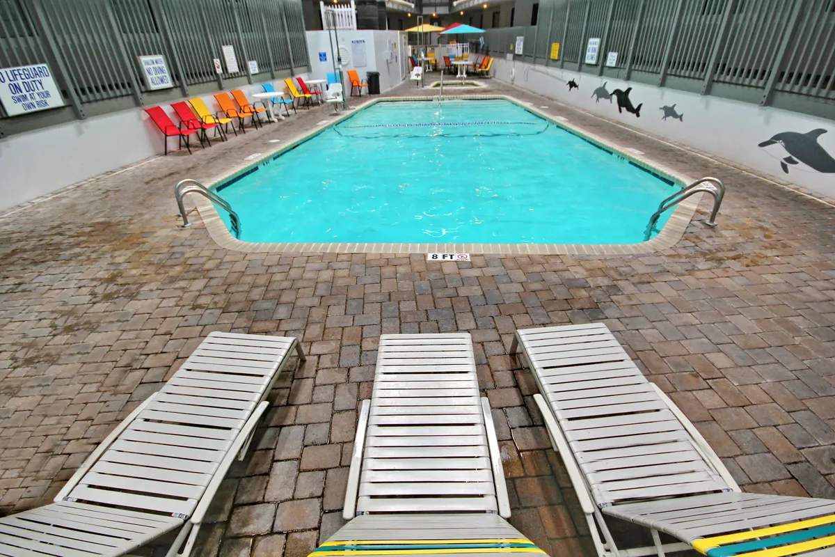 A vibrant outdoor pool scene featuring a large swimming pool surrounded by colorful lounge chairs and a prominent lifeguard station. The area is bright and lively, with plenty of space for sunbathing, swimming, and poolside activities, all under a sunny sky.
