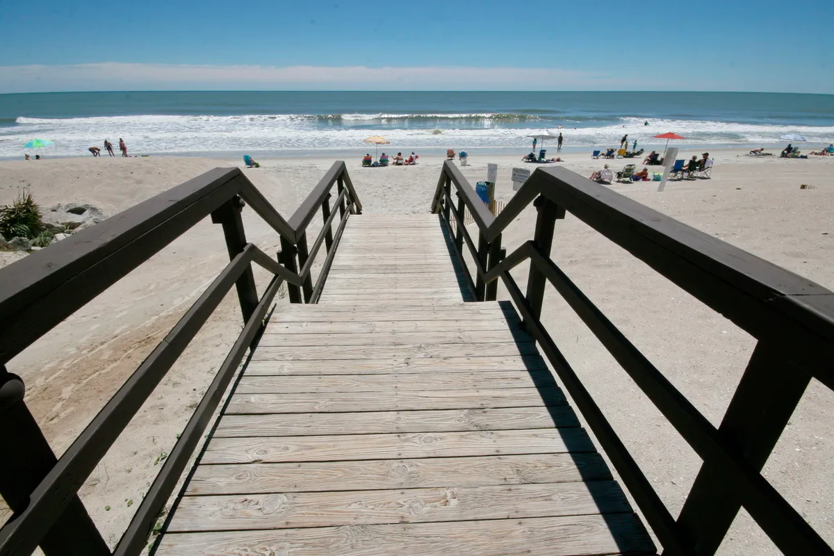 A scenic view of a wooden boardwalk leading to the ocean, with beachgoers enjoying sunbathing, swimming, and picnicking. The atmosphere is relaxed and inviting, with the waves and beach stretching out in the background.