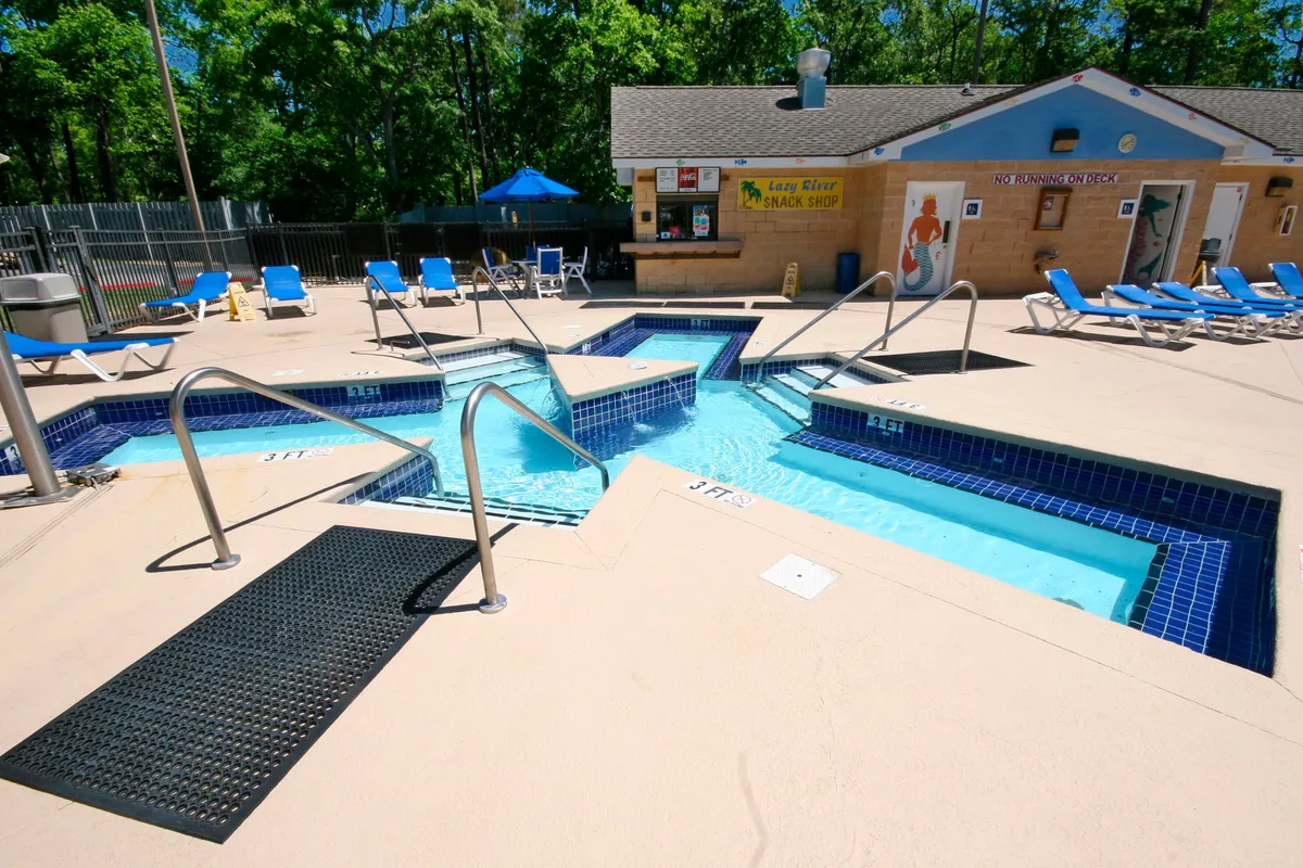 A lively pool scene with a snack shop, lounge chairs, handrails, and children playing in the water, while adults relax nearby. The area exudes a relaxed and cheerful mood, emphasizing both play and leisure.
