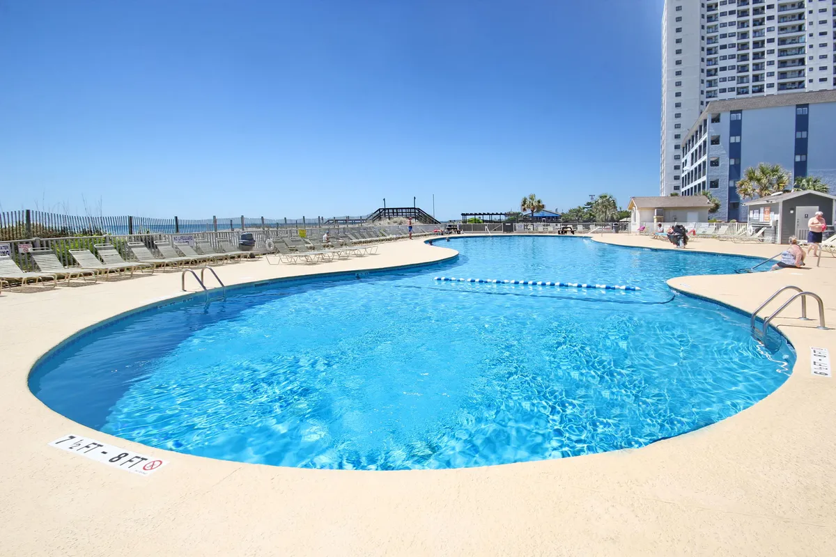 A sparkling outdoor swimming pool surrounded by comfortable lounge chairs and tall palm trees, with a high-rise resort building in the background. The scene is bright and inviting, suggesting both relaxation and social opportunities on a leisurely day.