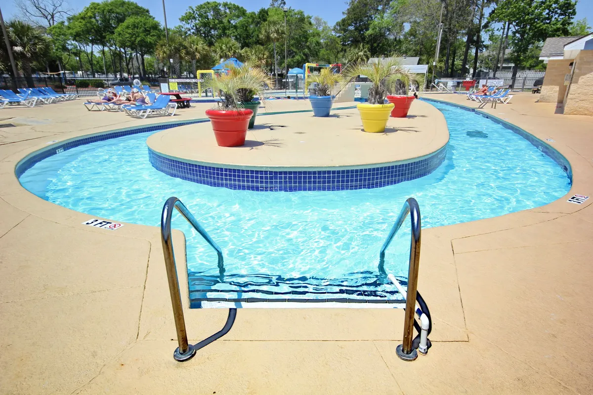 A vibrant swimming pool area enhanced by colorful planters and numerous poolside lounge chairs, creating an inviting space for both socializing and relaxation. The atmosphere is relaxed, capturing the essence of a fun-filled resort day.