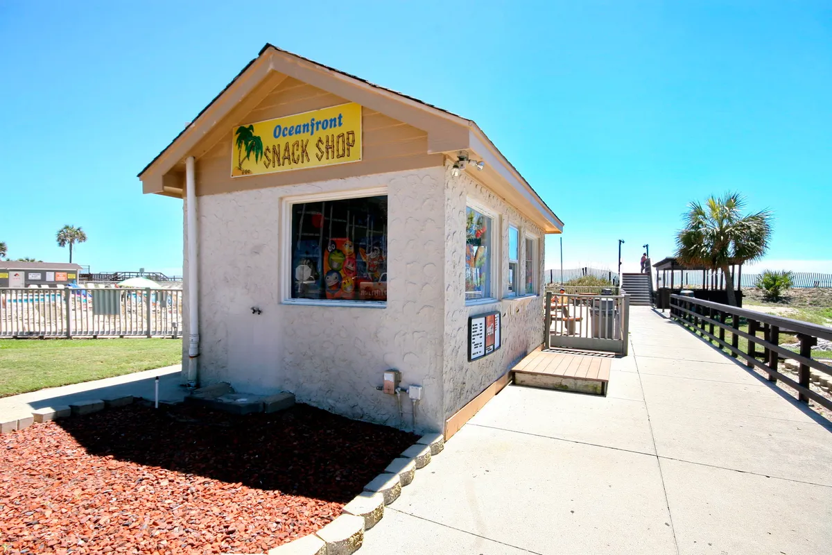 An inviting beachfront snack shop with colorful signage sits amid palm trees, with a walkway leading directly to the sandy beach. The scene radiates a casual, vacation vibe and highlights the easy access to both refreshments and the shoreline.