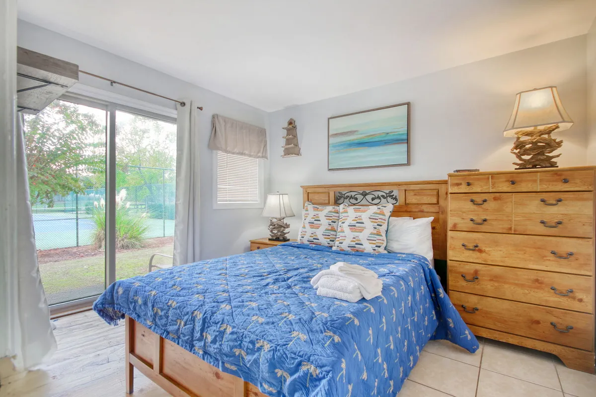 A bright, inviting bedroom with a plush bed, nightstand with lamp, dresser, and a window offering a view of the tennis courts and lush greenery beyond. Decorative artwork adorns the wall, and soft natural light fills the space, creating a tranquil retreat.
