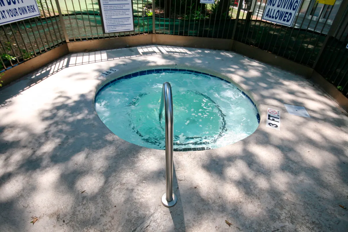 A tranquil hot tub set amidst lush greenery, complete with a handrail for easy access. The sun glistens on the water’s surface, inviting guests to unwind and enjoy peaceful moments outdoors.