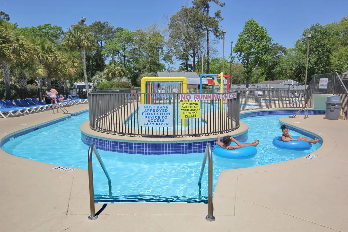 A vibrant exterior pool area featuring a winding lazy river, a colorful splash area for kids, and plenty of lounge chairs set against lush greenery. Children are seen playing in the water while families relax on the poolside, and the playful design of the water features creates an inviting, joyful atmosphere.